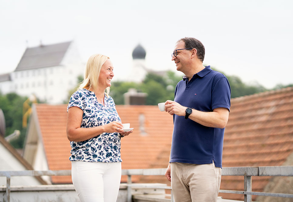 Zwei Personen stehen auf einer Terasse mit Gebäuden im Hintergrund.